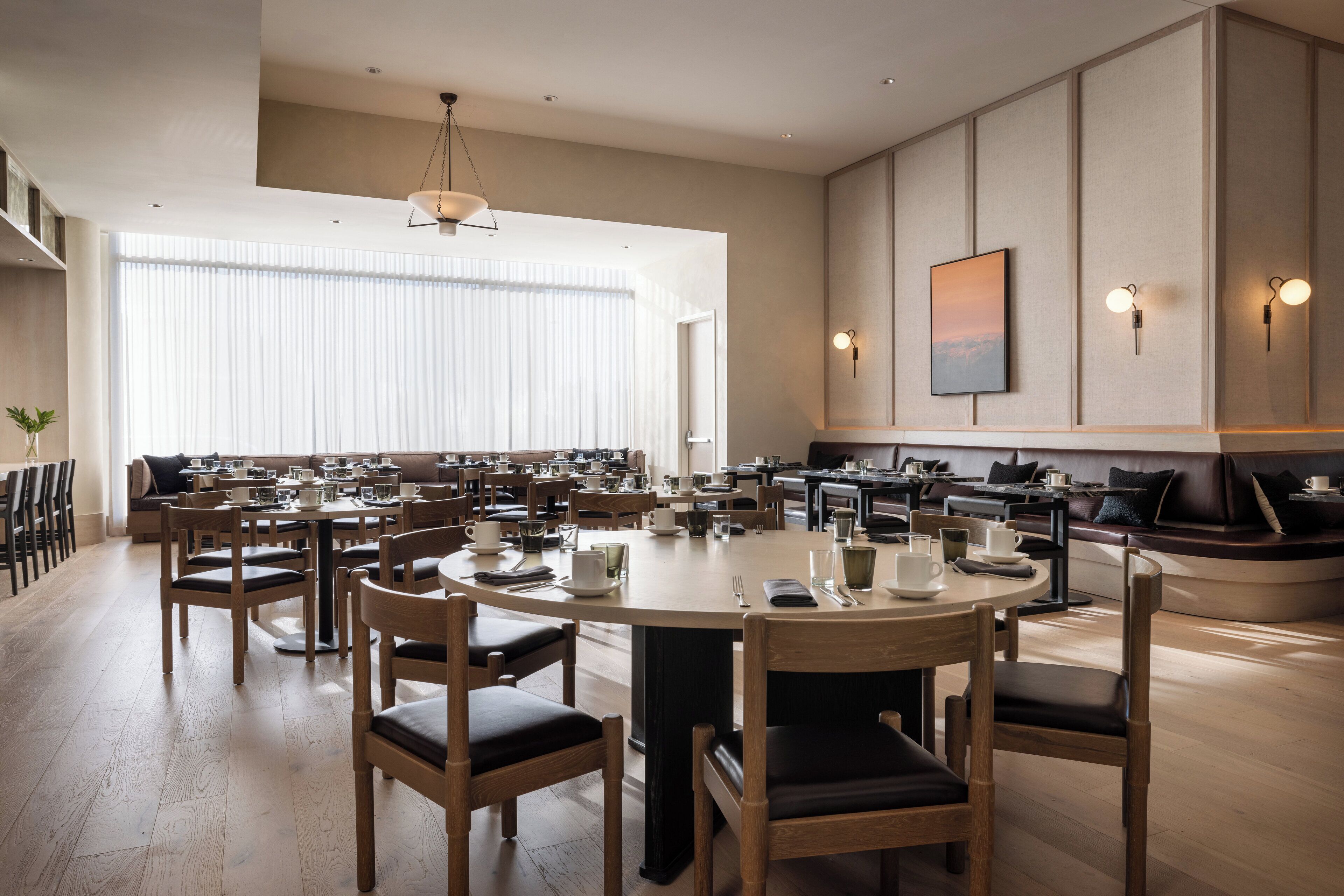 A spacious dining area with multiple round tables and chairs, a large window with blinds, and wall-mounted lighting fixtures.