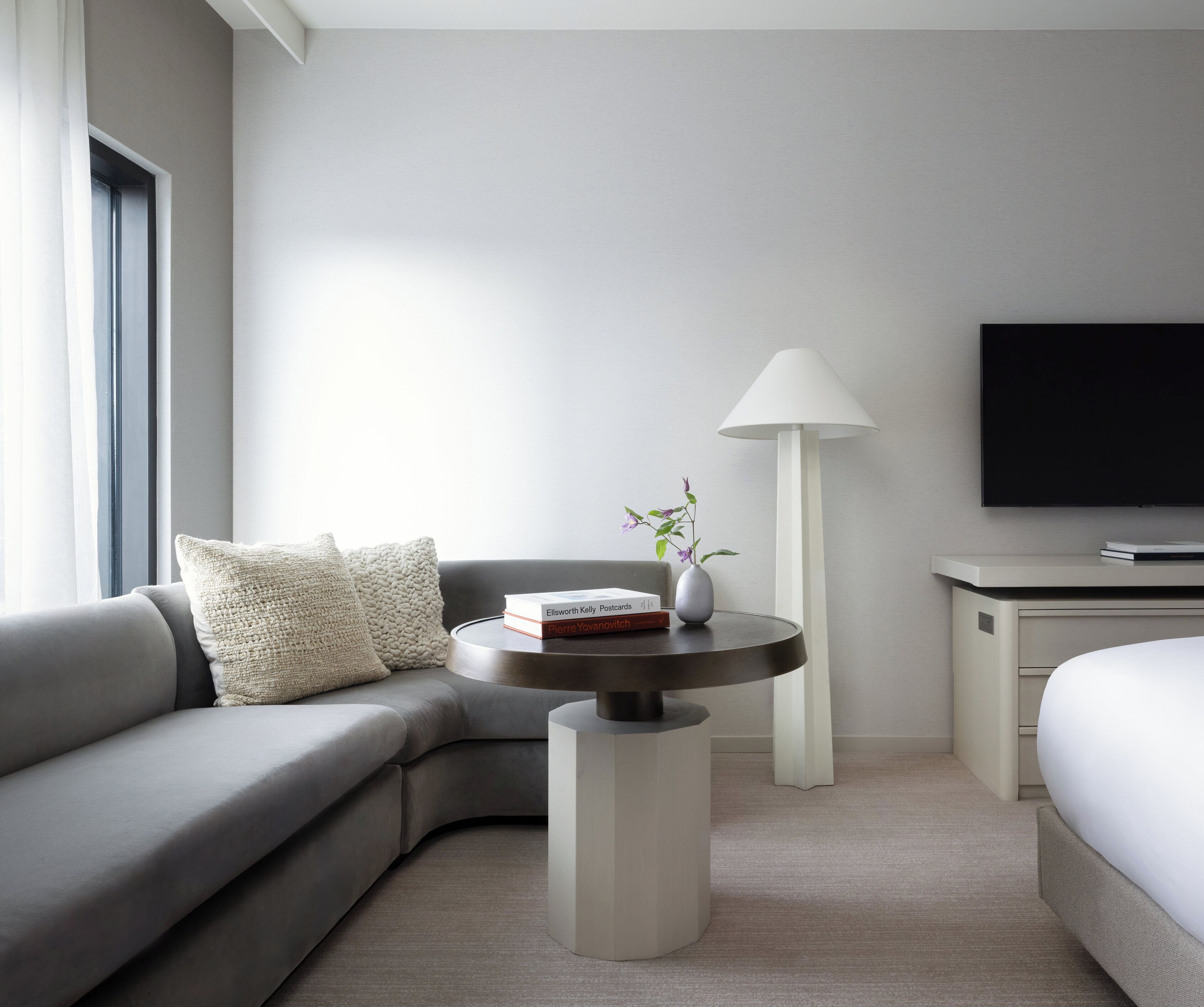 A modern hotel room with a grey sofa, a small round table with a lamp, a vase with flowers, and a flat-screen TV on a stand.