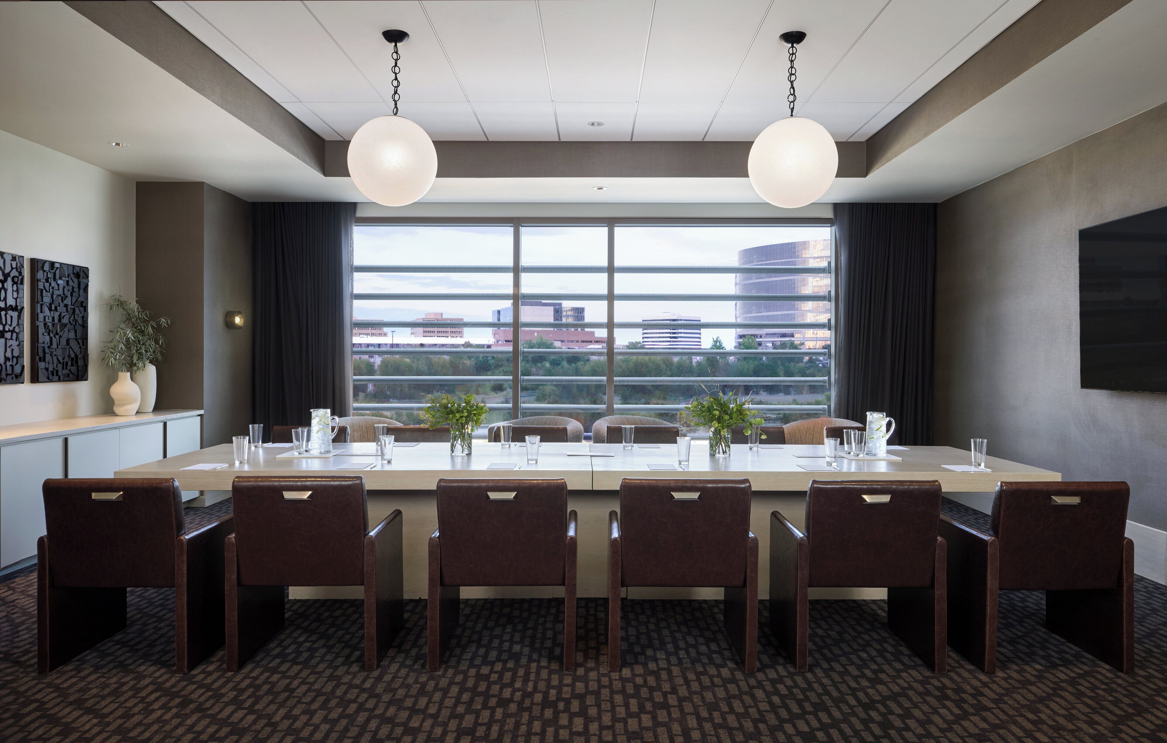 A conference room with a long table, six chairs, two pendant lights, a large window with a city view, and a plant on the side.