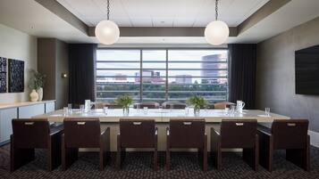 A conference room with a long table, six chairs, two pendant lights, a large window with a city view, and a plant on the side.