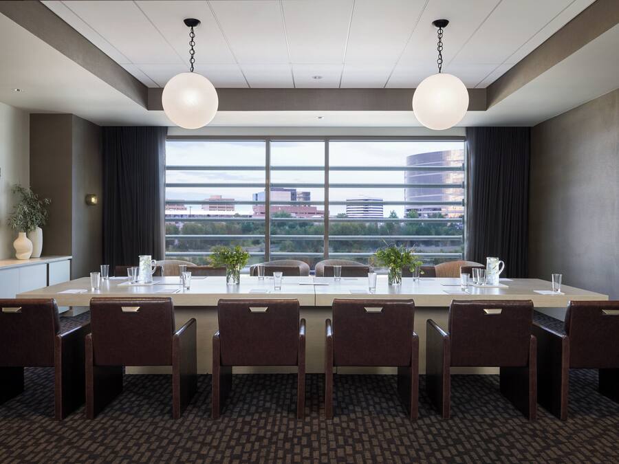 A conference room with a long table, six chairs, two pendant lights, a large window with a city view, and a plant on the side.
