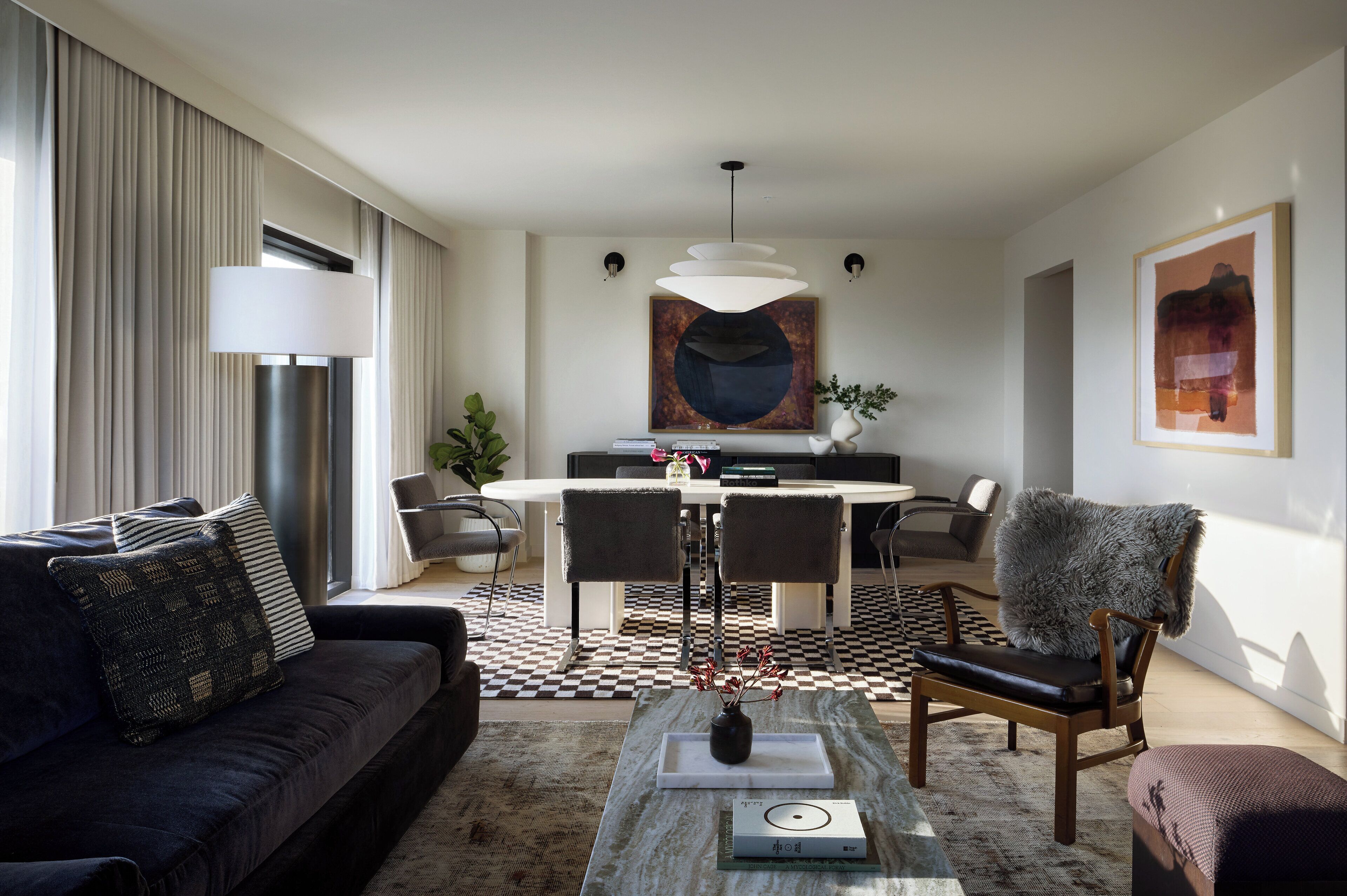 A modern living room with a sofa, coffee table, and chairs. There is a dining area with a table and chairs. The room has large windows with curtains.