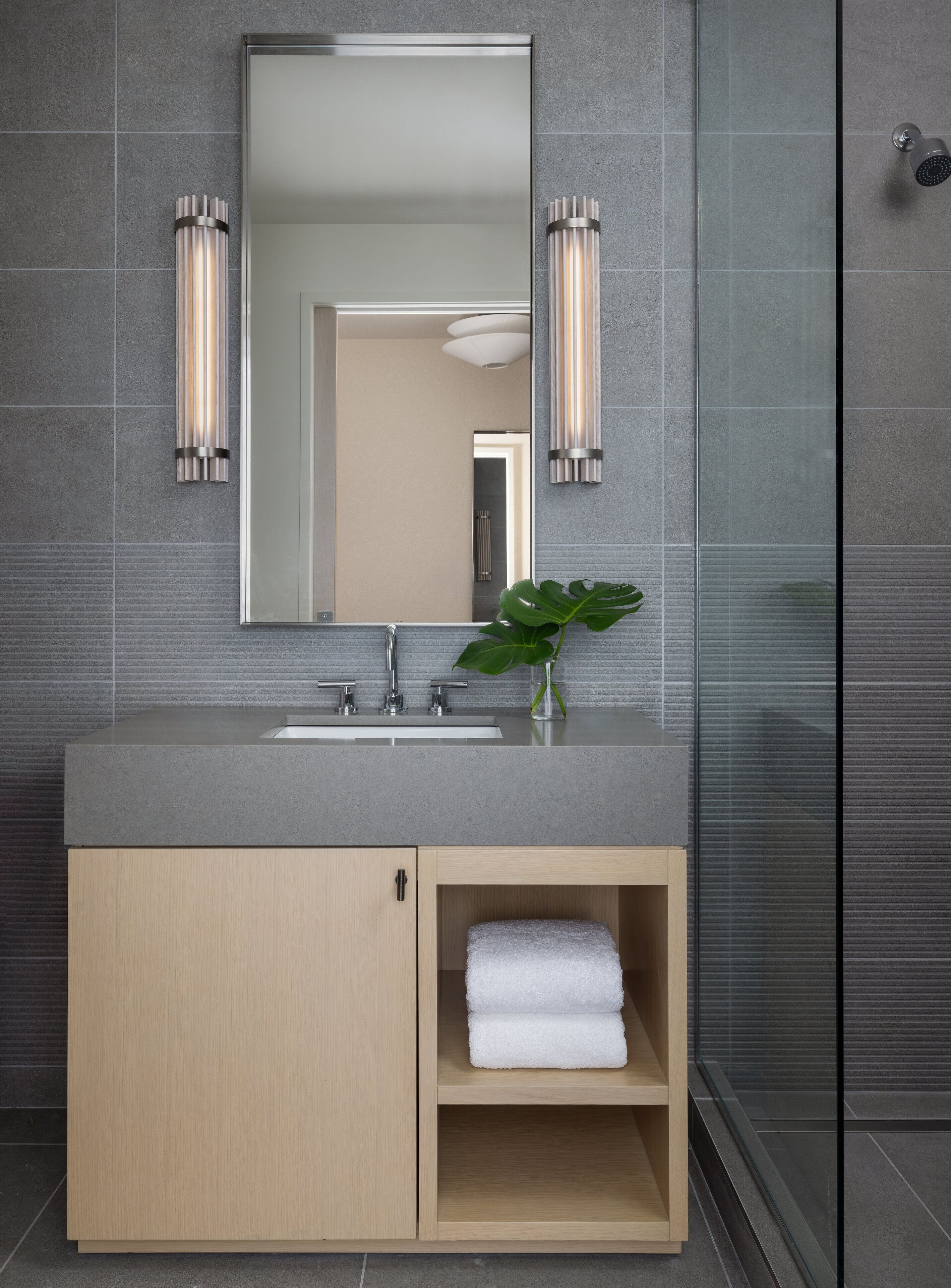 Un baño moderno con pared de azulejos grises, un espejo grande, un lavamanos con dos grifos y una planta en un jarrón.