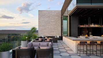 A rooftop bar with outdoor seating, a bar counter, and a large window overlooking the city.