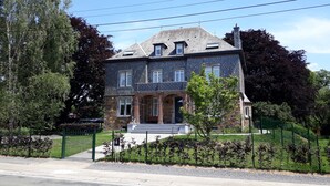 A two-story stone house with a gabled roof, a front porch, and a fenced yard. - L'Albizia Maison d'Hôtes (Libin)