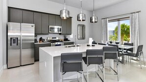 Een moderne keuken met roestvrijstalen apparatuur, een centraal kookeiland met barkrukken, hanglampen en uitzicht op de omgeving door grote ramen.