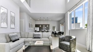 A modern living room with a grey sofa, a glass coffee table, and a grey armchair. There is a kitchen in the background with pendant lighting.