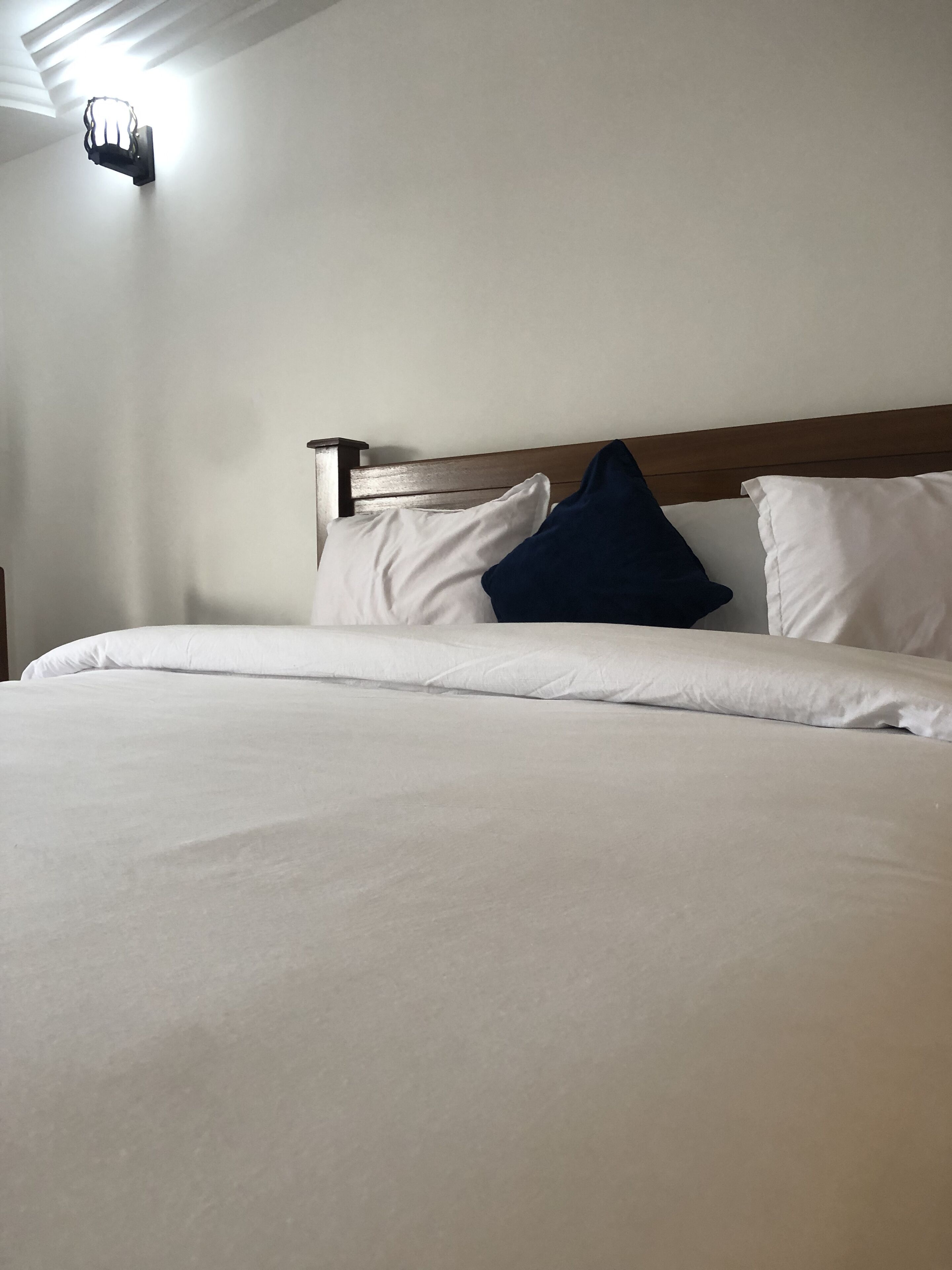 A neatly made bed with white and dark blue pillows, a wooden headboard, and a wall-mounted light fixture.