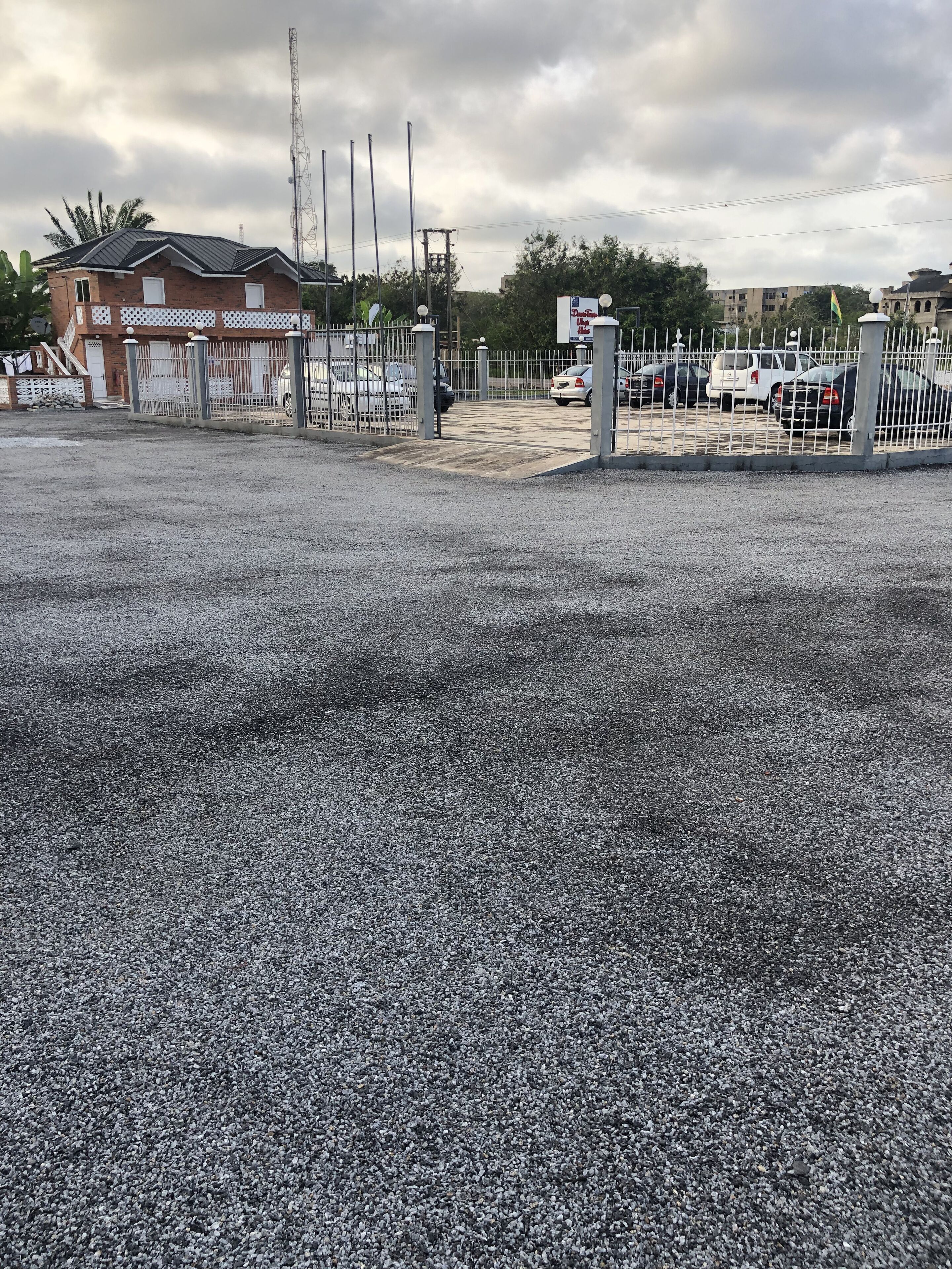 Un área de estacionamiento con superficie de grava, entrada protegida por una reja y un edificio de fachada de ladrillo.