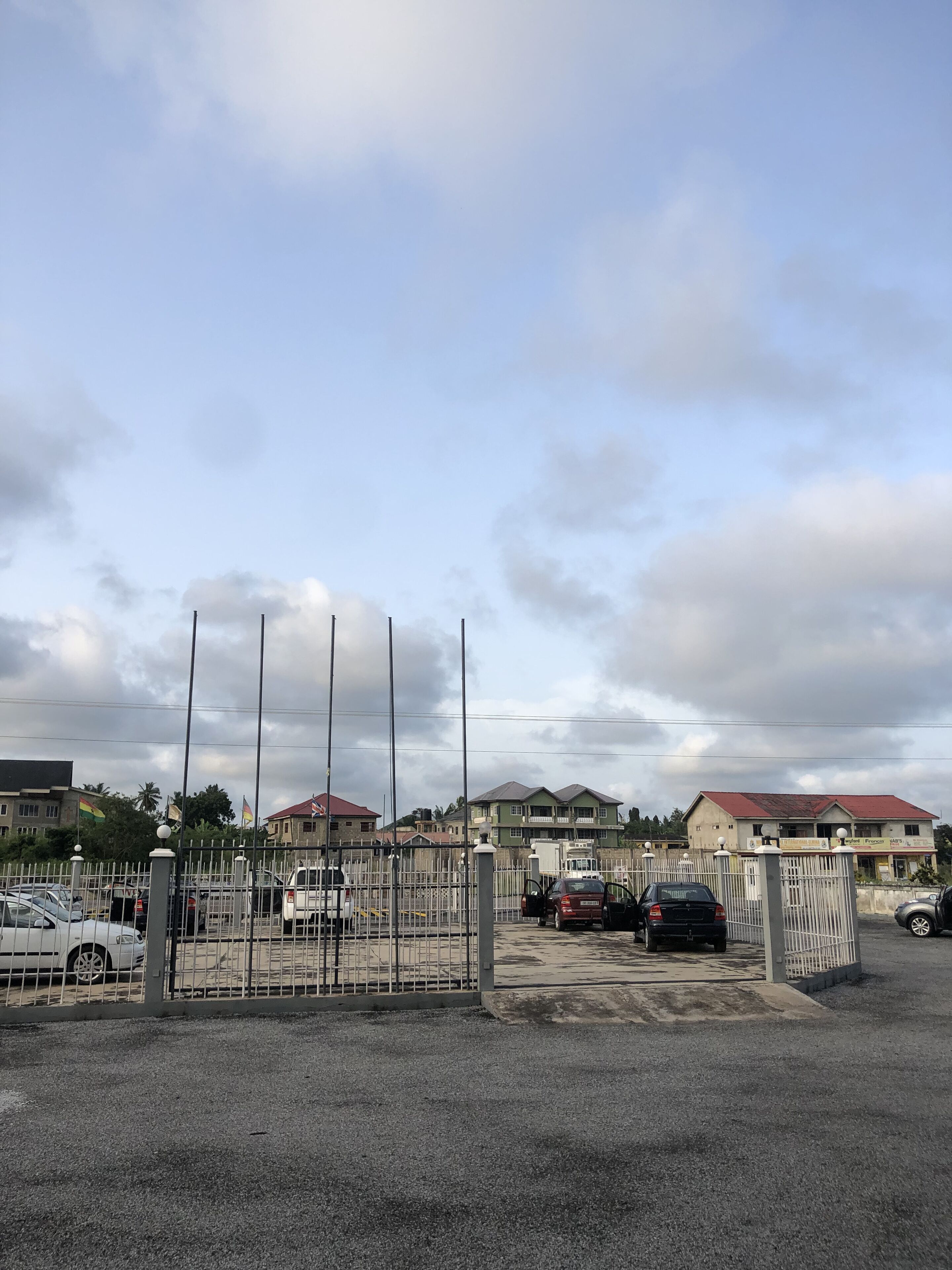 Area di parcheggio recintata con diverse auto, cielo sereno e edifici sullo sfondo.