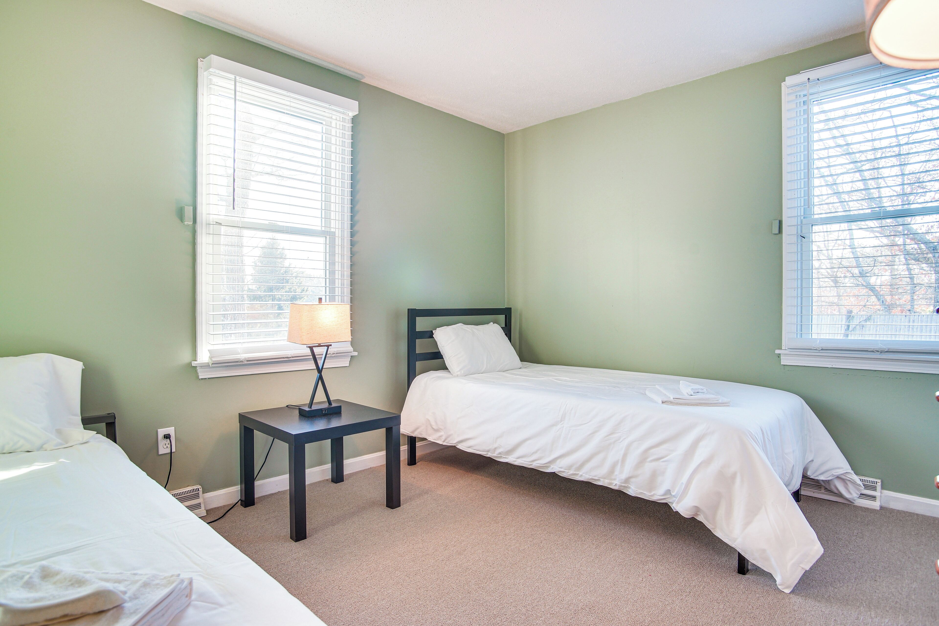 Une chambre avec deux lits, une table de chevet, une lampe et deux fenêtres équipées de stores.