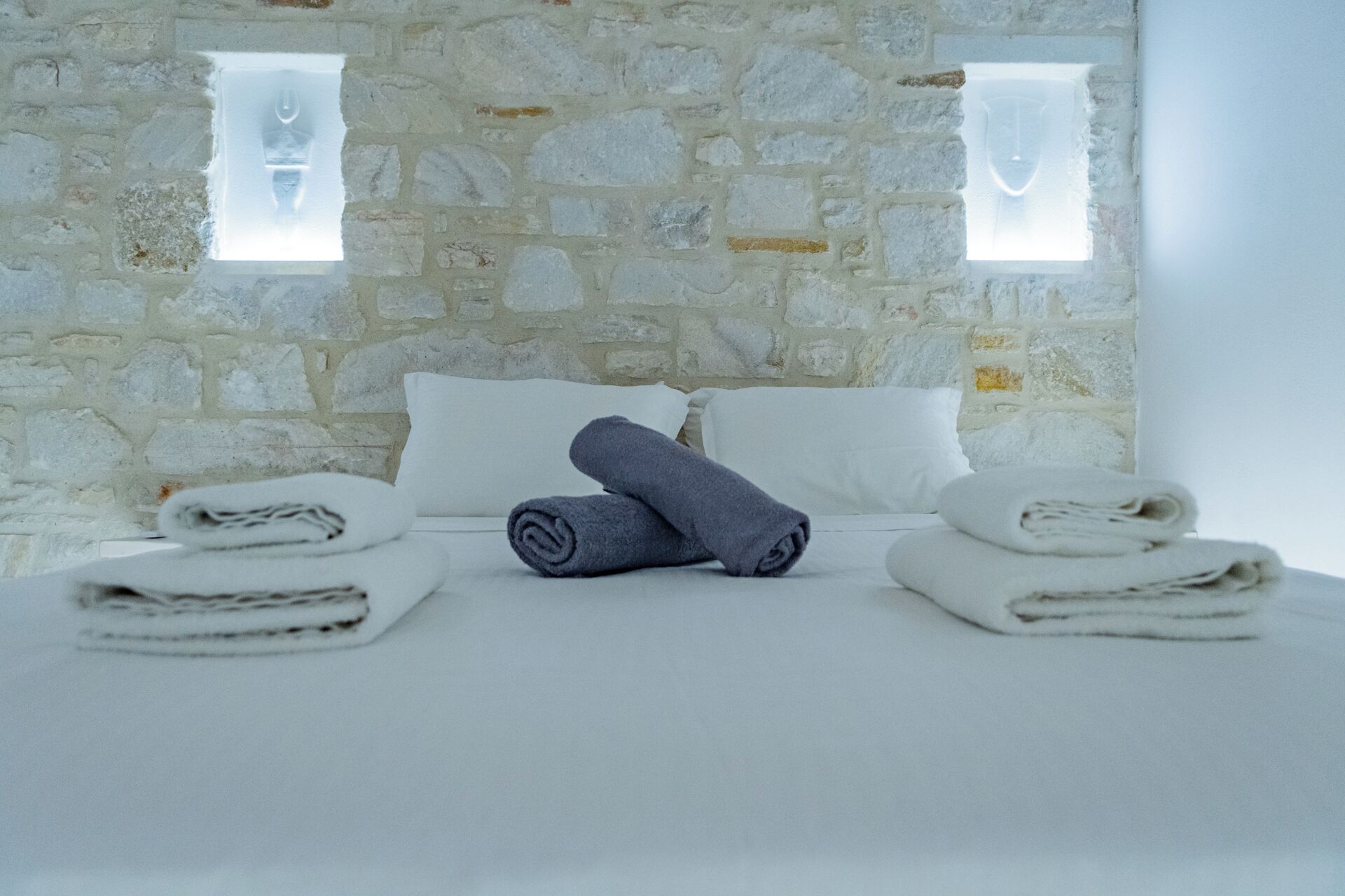 A room with a stone wall, a bed with white linens, and two windows with frosted glass.
