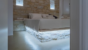 A bedroom with a stone wall, a bed with white linens, a small window, and a mirror. - Aegean Feelings (Paros)