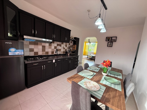 A modern kitchen with dark cabinets, a stainless steel range hood, and a tiled backsplash. There is a dining area with a wooden table set for four, featuring green placemats and a vase with flowers.