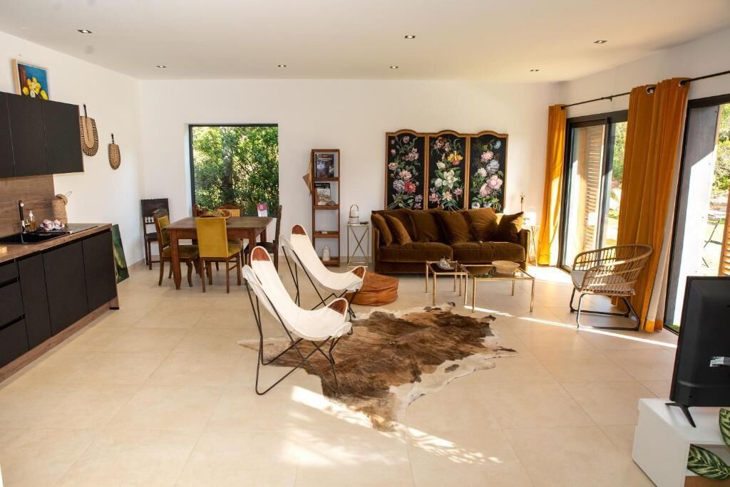 A modern living room with a sofa, chairs, a coffee table, and a rug. There is a dining area with a table and chairs, and a kitchen area with cabinets and a counter.