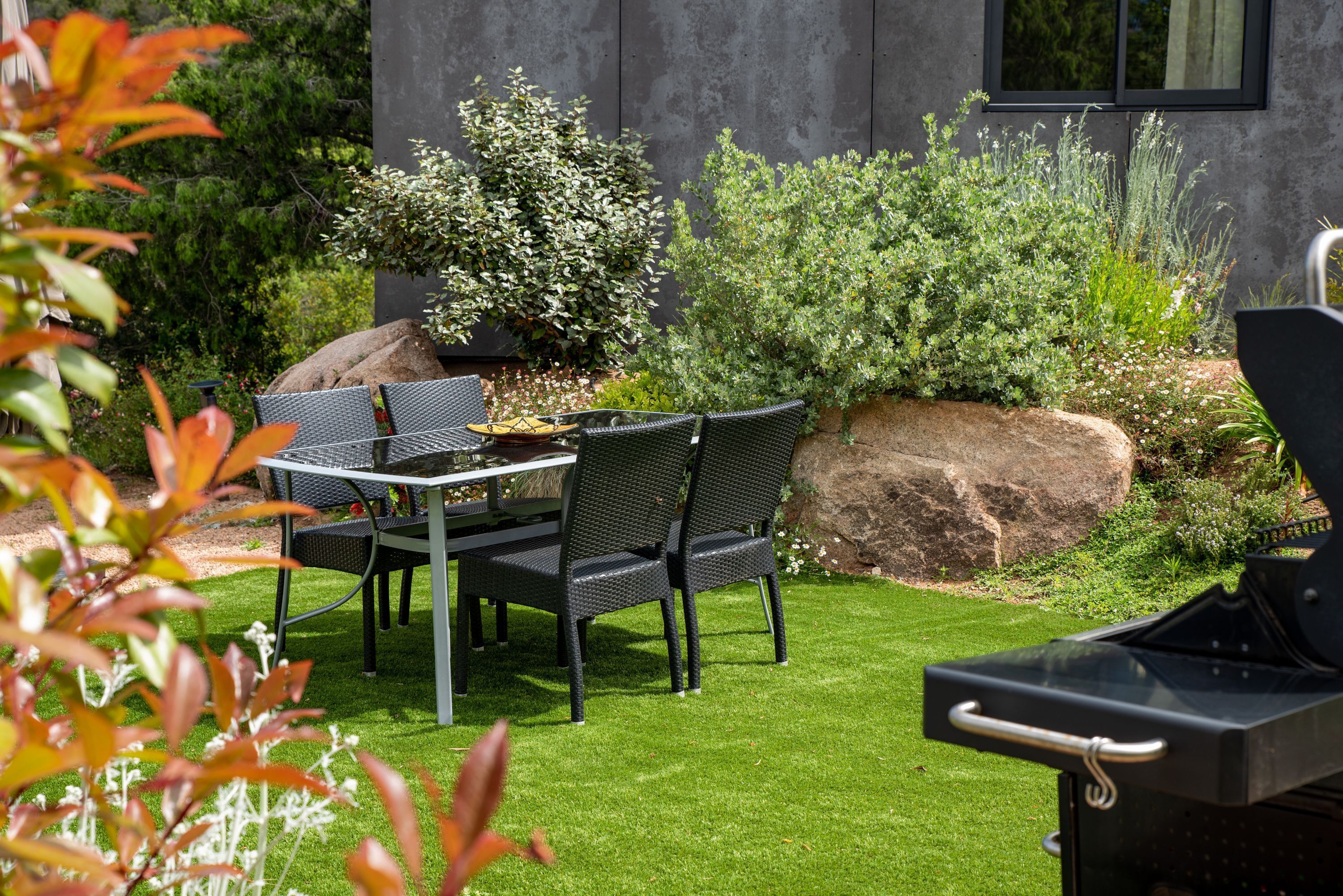 An outdoor patio with a table and chairs, surrounded by greenery and a barbecue grill.