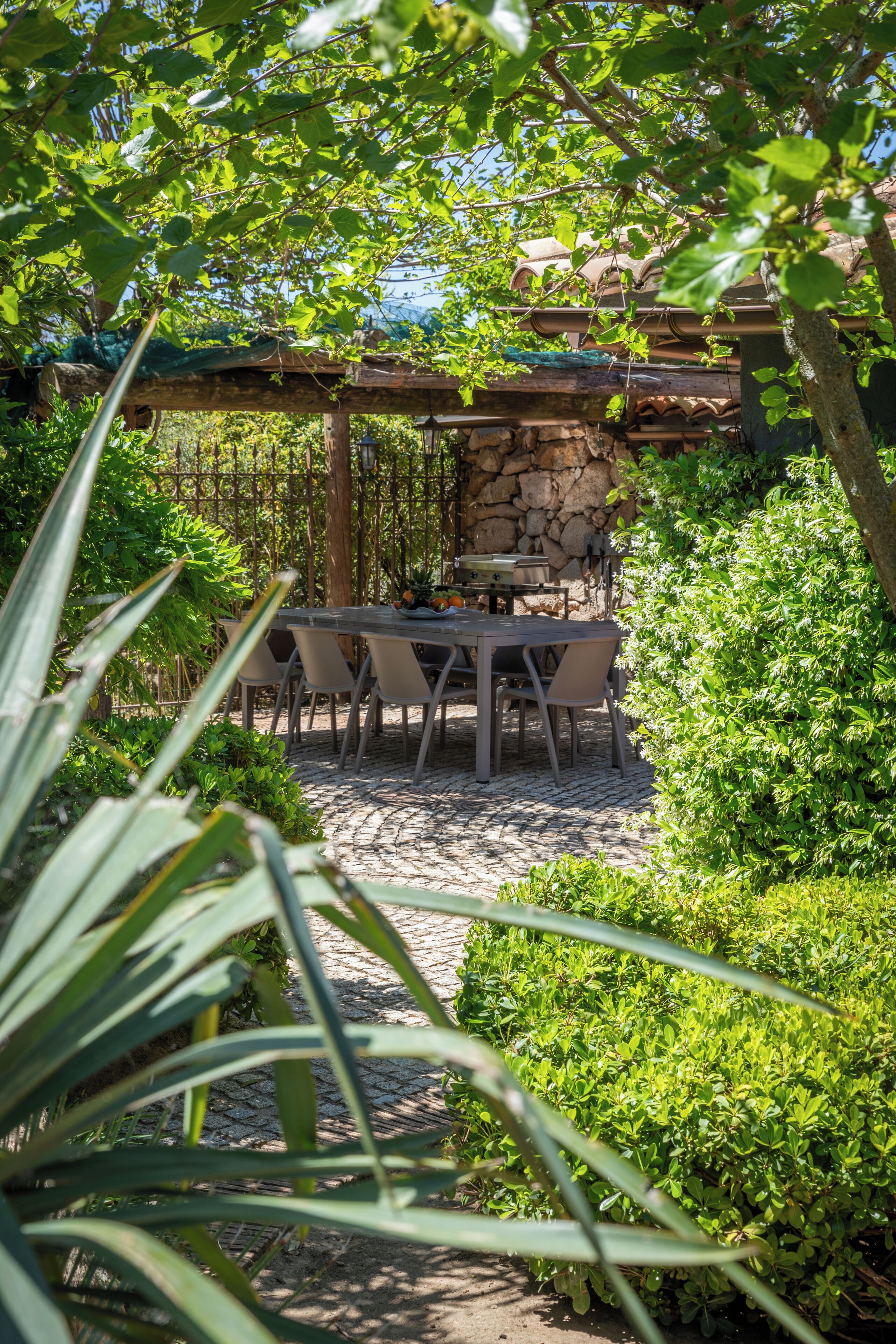 Un espace patio avec une table et des chaises en pierre, entouré d’une végétation luxuriante et d’un mur de pierre.