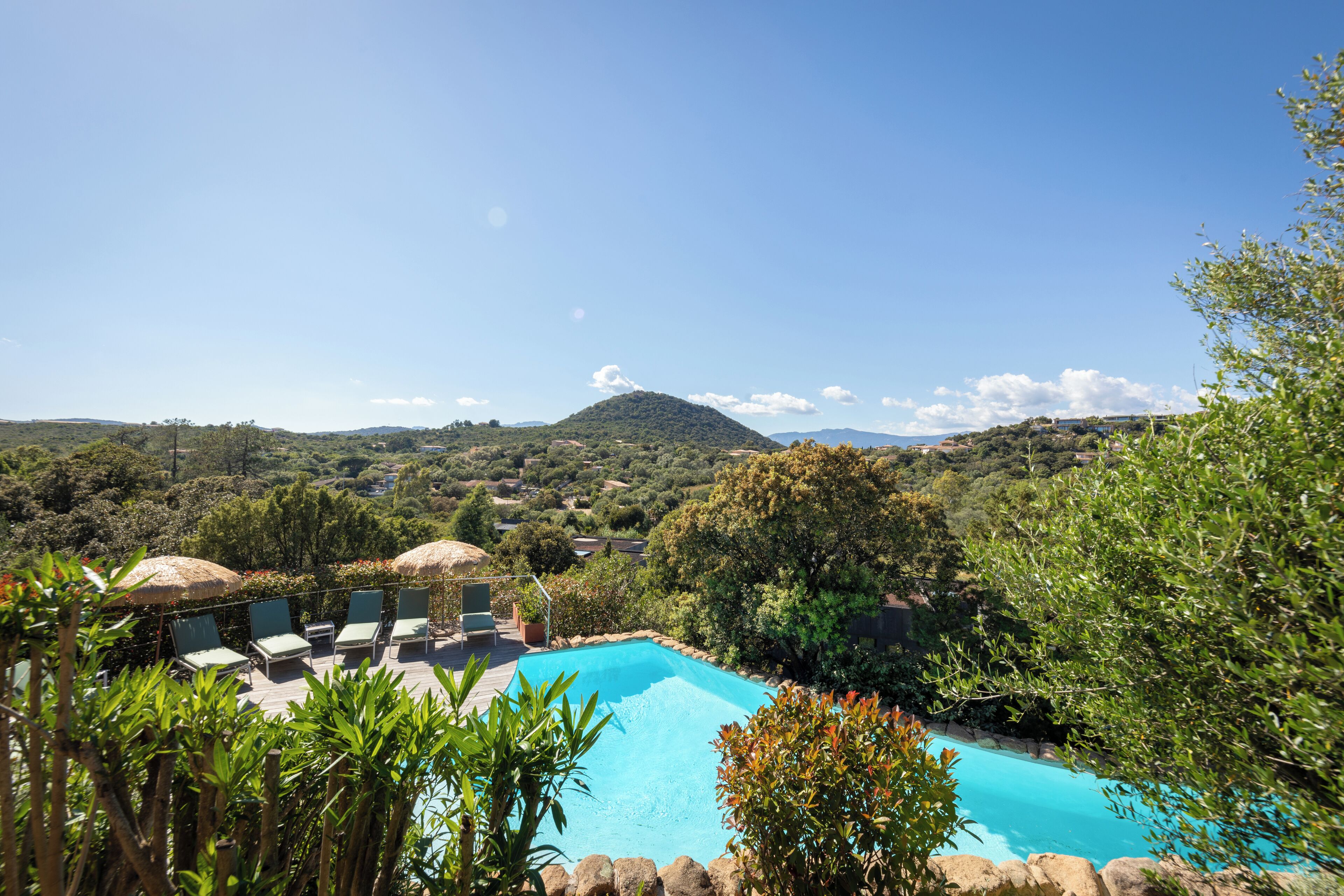 A pool with lounge chairs and umbrellas, surrounded by greenery and hills.