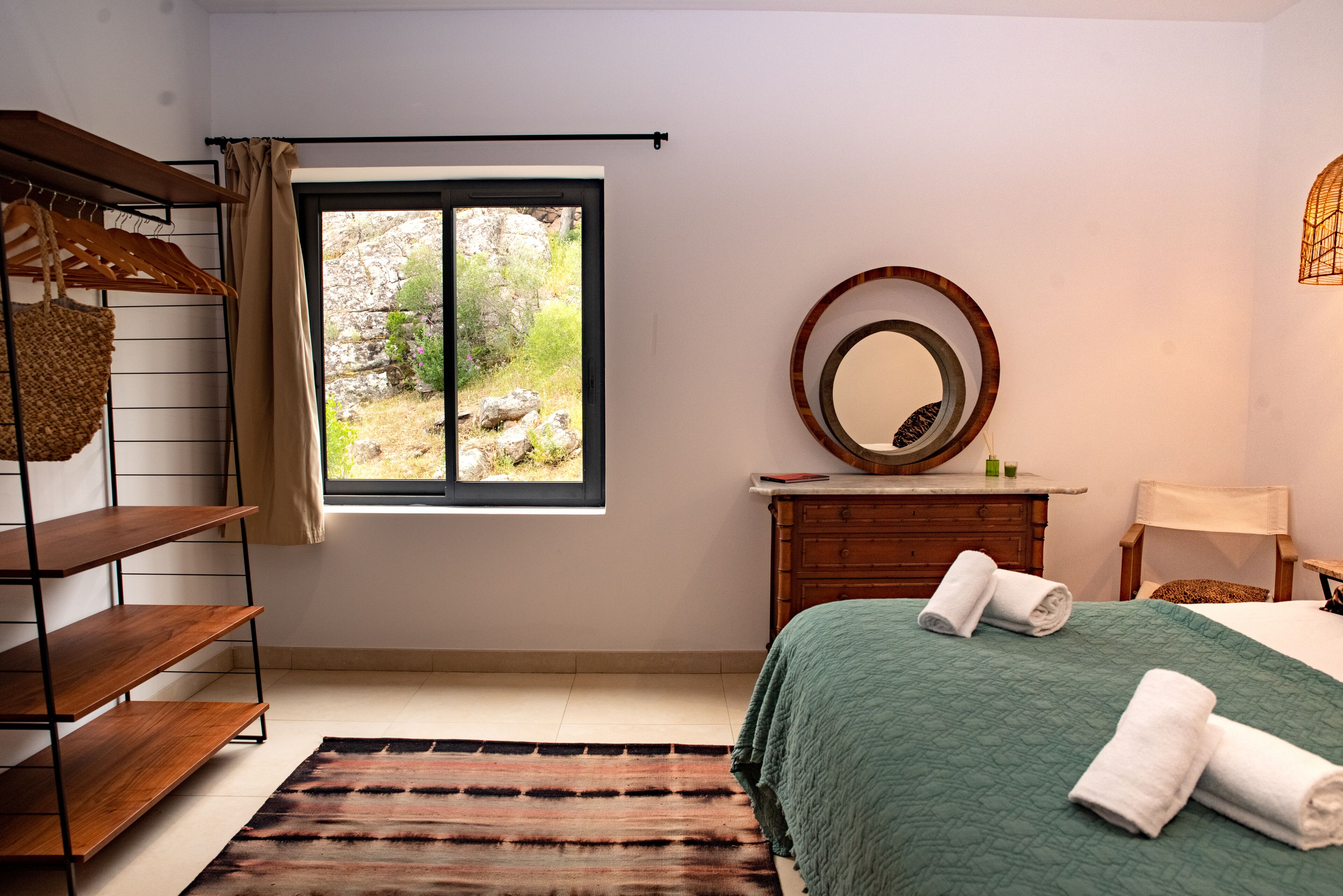 A bedroom with a bed, a wooden dresser, a mirror, a window with a view of greenery, and a shelf with towels.