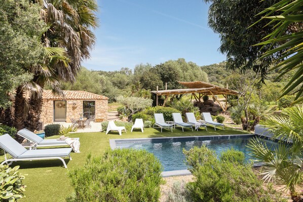 Un espace piscine avec des chaises longues, un bâtiment en pierre et une pergola en bois.