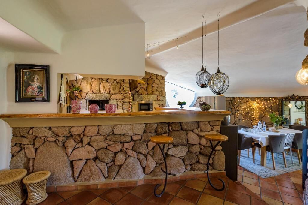 Zona de bar de piedra con chimenea rústica, luces colgantes y comedor con mesa preparada para dos.