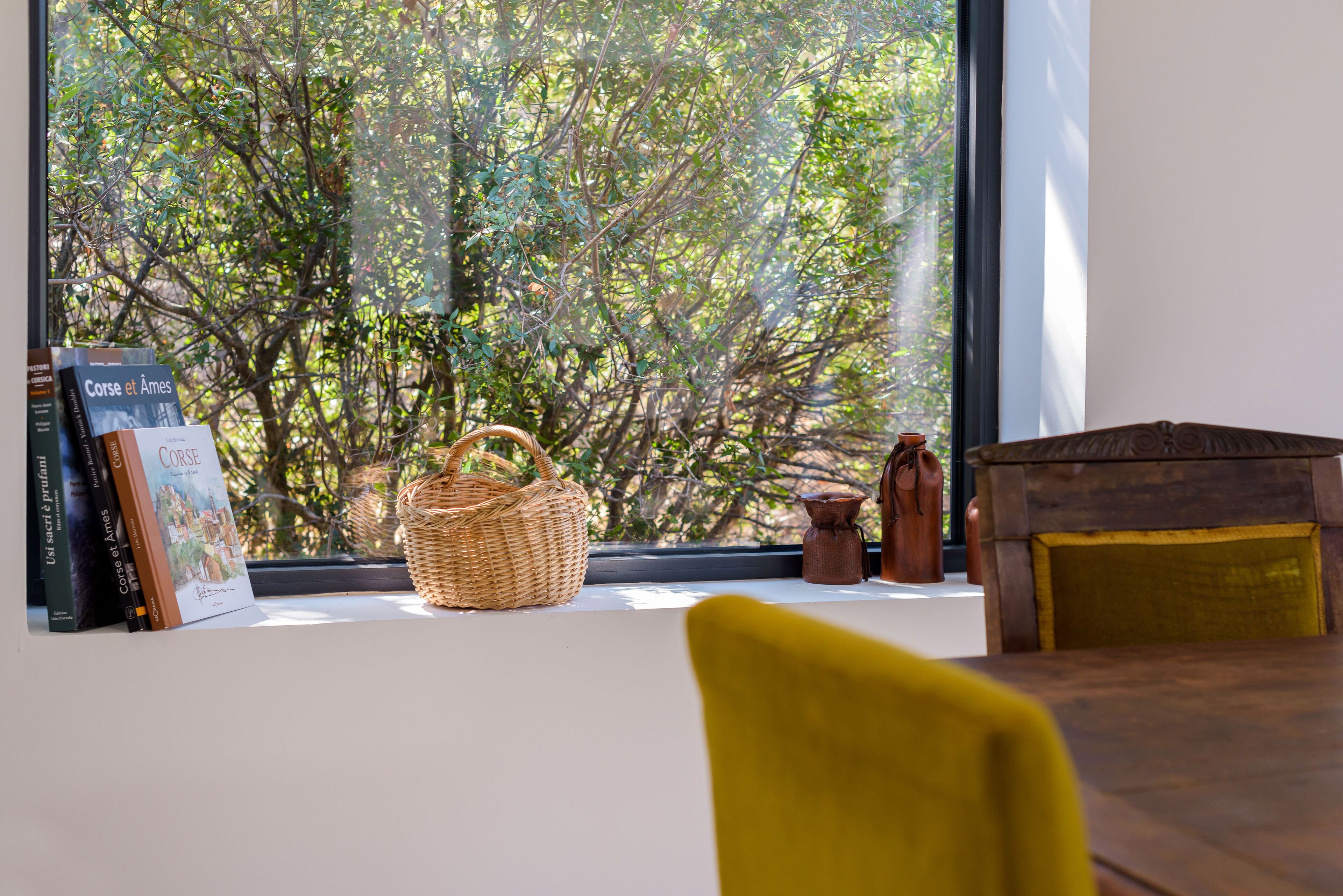 A room with a large window overlooking a tree, a wicker basket, a small wooden chest, and a yellow chair.