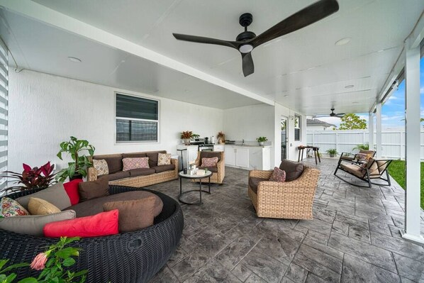 A covered patio area with wicker furniture, a ceiling fan, and a kitchen in the background.
