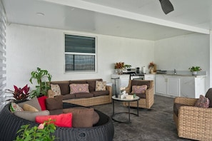 A covered outdoor living area with wicker furniture, a kitchen in the background, and potted plants.