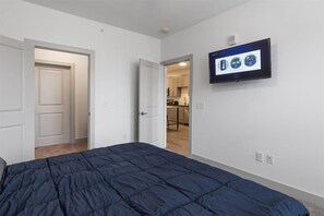 Ein Schlafzimmer mit einem Bett, einem an der Wand montierten Fernseher und einer zur Küche führenden Tür.