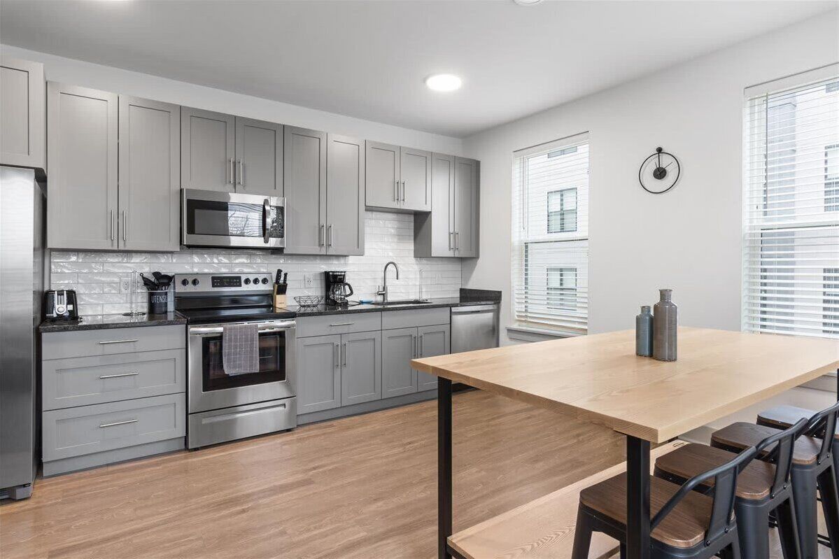 Una cocina moderna con armarios grises, electrodomésticos de acero inoxidable y una mesa de comedor de madera con taburetes de bar.