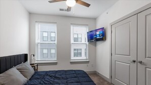 Quarto com cama, ventilador de teto, TV fixada na parede e uma porta.