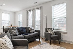 A modern living room with a grey sofa, armchair, and a coffee table. There are large windows with blinds, a floor lamp, and a rug on the floor. - Experience Cincinnati! Walkable Modern Condo (Cincinnati)