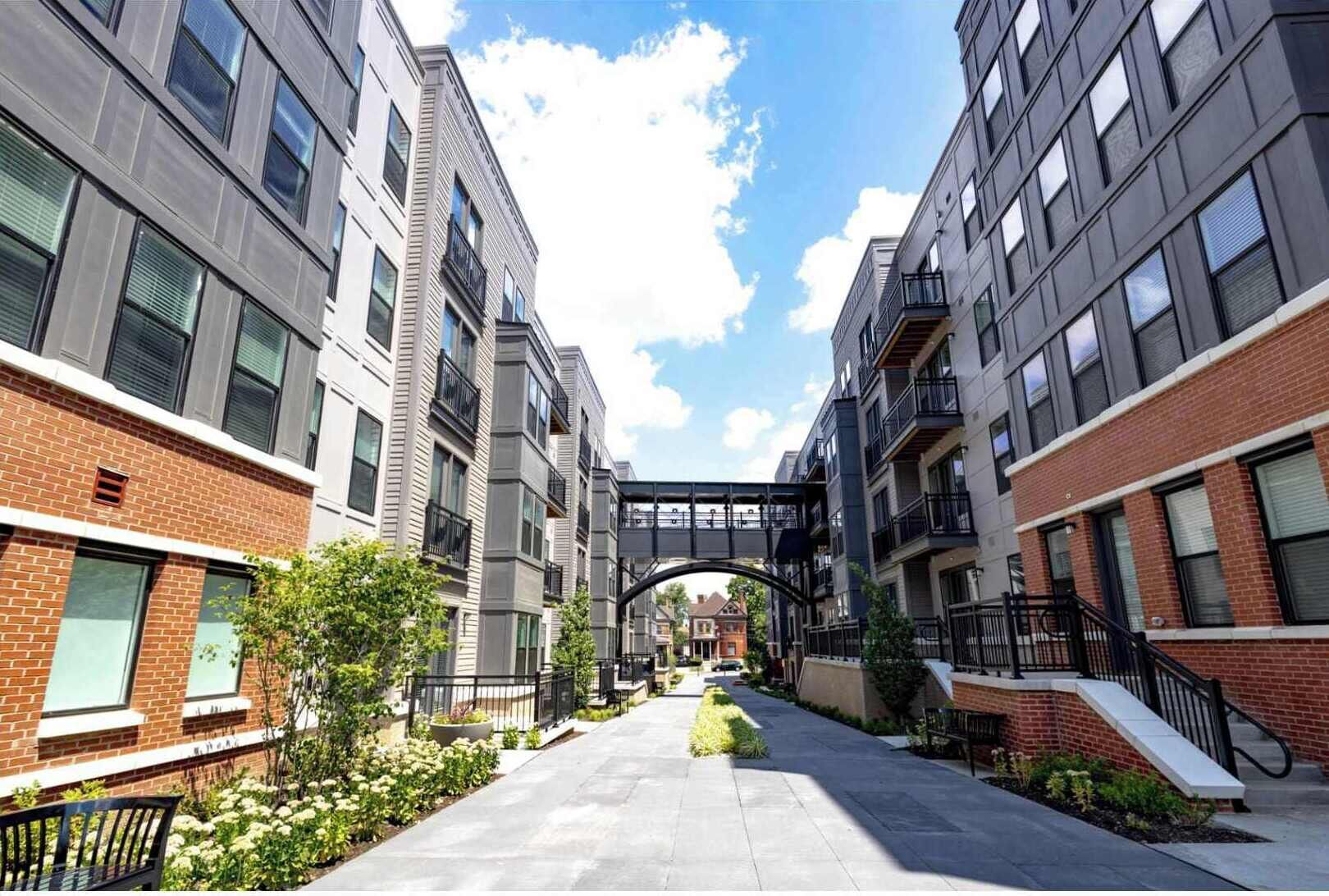 A multi-story residential building with a central pedestrian bridge, surrounded by greenery and a clear sky.