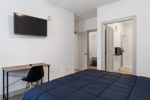 A bedroom with a bed, a desk, a chair, a TV, and a bathroom visible through an open door.