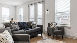 A modern living room with a grey sofa, armchair, and a coffee table. There are large windows with blinds, a floor lamp, and a rug on the floor. - Explore Cincinnati’s Best from a Chic Condo (Cincinnati)