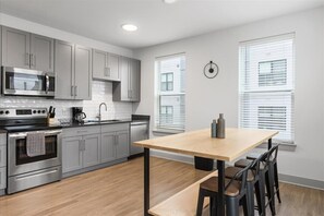 Eine moderne Küche mit grauen Schränken, einer Edelstahl-Mikrowelle und einem Backofen. Ein Holztisch mit Stühlen und eine Uhr an der Wand sind ebenfalls vorhanden.