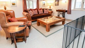 Een gezellige woonkamer met een leren fauteuil, een bruine bank en een houten salontafel. Er zijn twee ramen met gordijnen, een lamp en een lijst met een foto aan de muur.