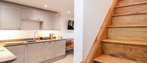 A modern kitchen with light-colored cabinets, a wooden staircase, and a flat-screen TV mounted on the wall.