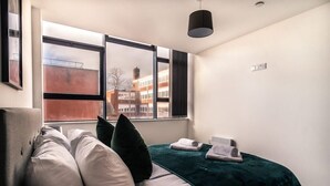 Moderni hotellihuone, jossa on suuri ikkuna, valkoisilla ja vihreillä tekstiileillä varustettu sänky, musta lamppu ja näkymä ulkona oleviin rakennuksiin.