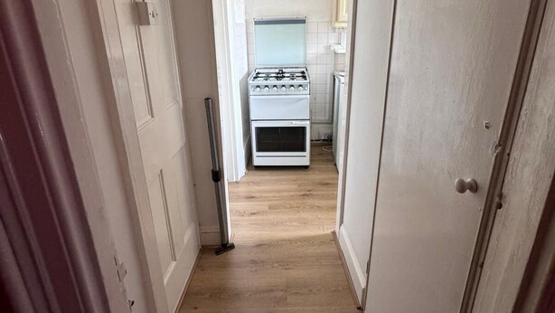 A narrow hallway with wooden flooring leading to a kitchen area with a gas stove and a refrigerator. - Walton Apartment (London)