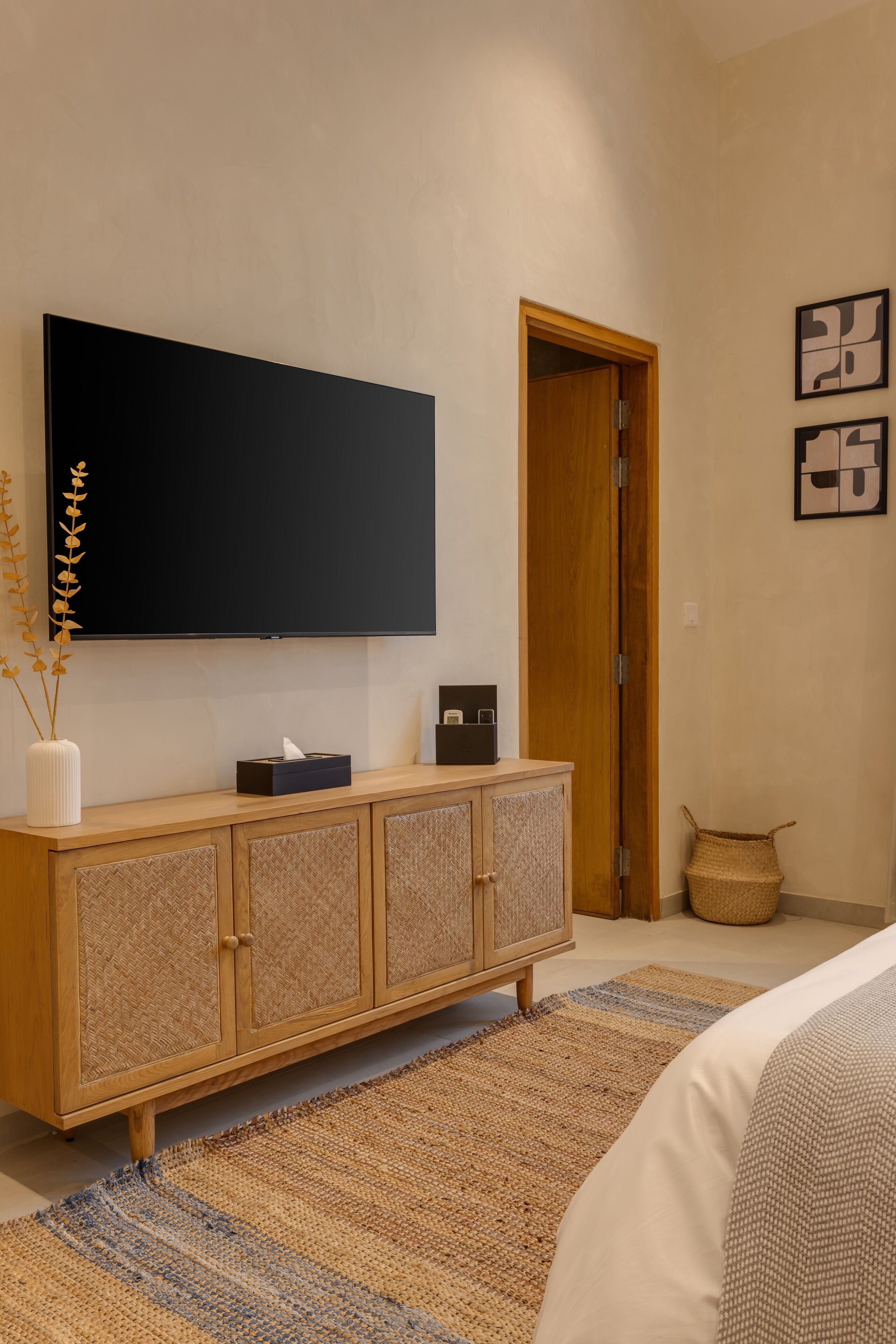 A modern bedroom with a large flat-screen TV mounted on the wall, a wooden cabinet with a woven texture, a woven rug, and a woven basket on the floor.