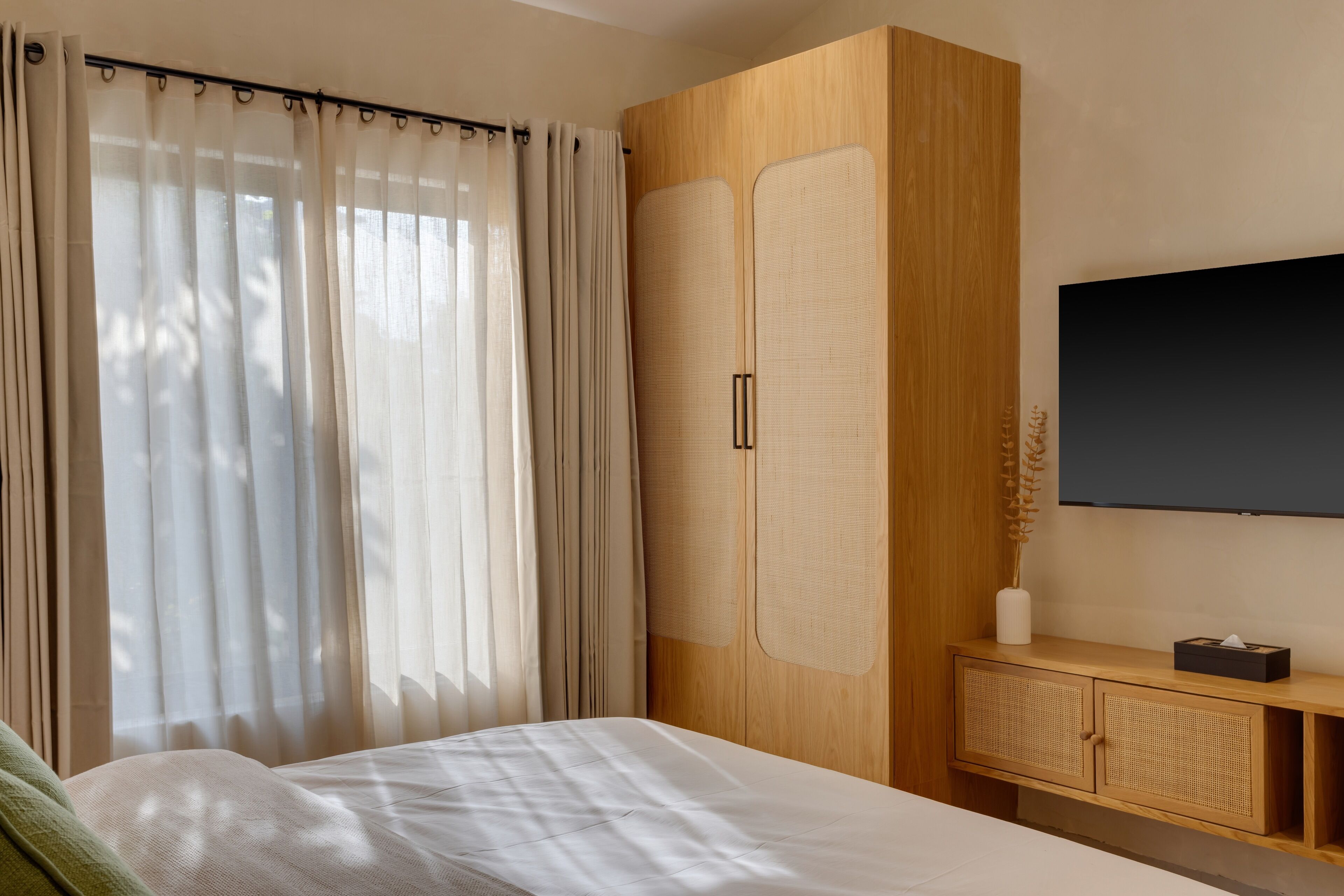 A bedroom with a bed, a wooden wardrobe, a TV, and a bedside table with a vase.