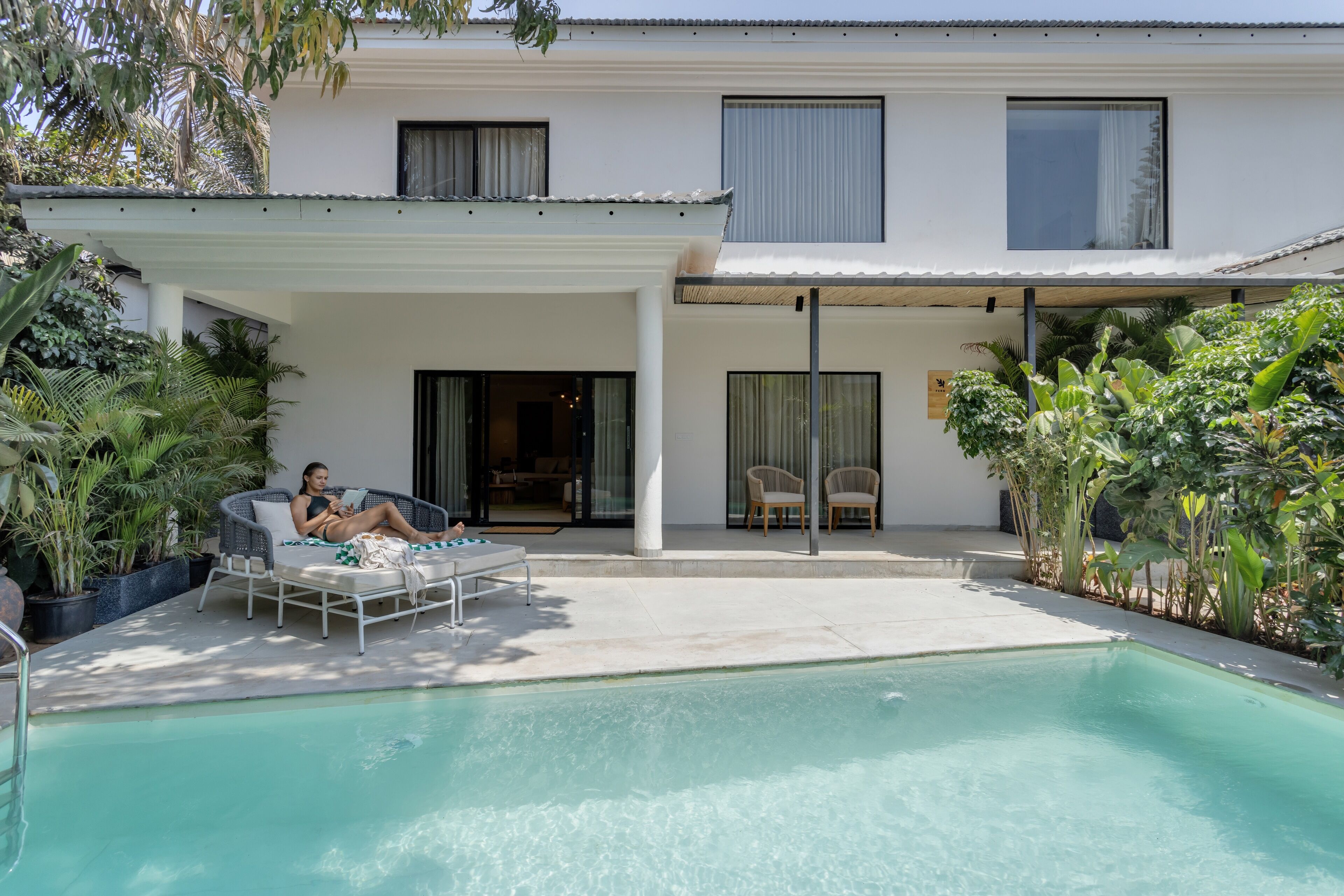 Zona de piscina con sillones de descanso, un porche cubierto y un edificio con amplias ventanas y un balcón.