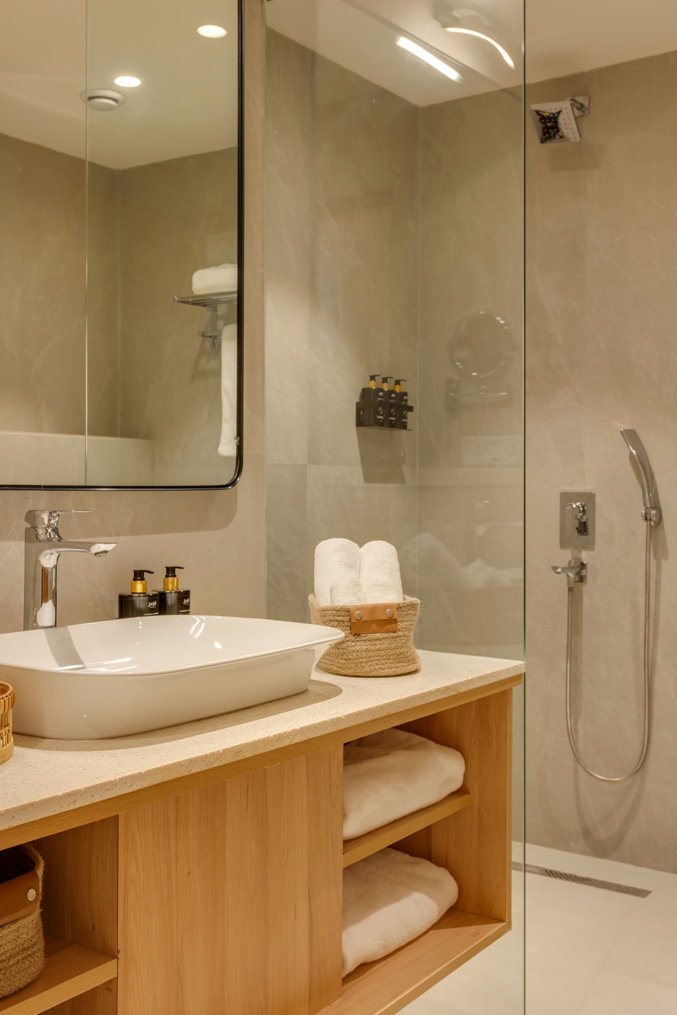 Un baño moderno con un espejo grande, un lavamanos blanco, estantes de madera y una zona de ducha con mampara de vidrio.