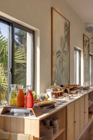 Um buffet de café da manhã com diversas opções de alimentos e bebidas, uma grande janela com vista para palmeiras e uma pintura com temática tropical na parede.