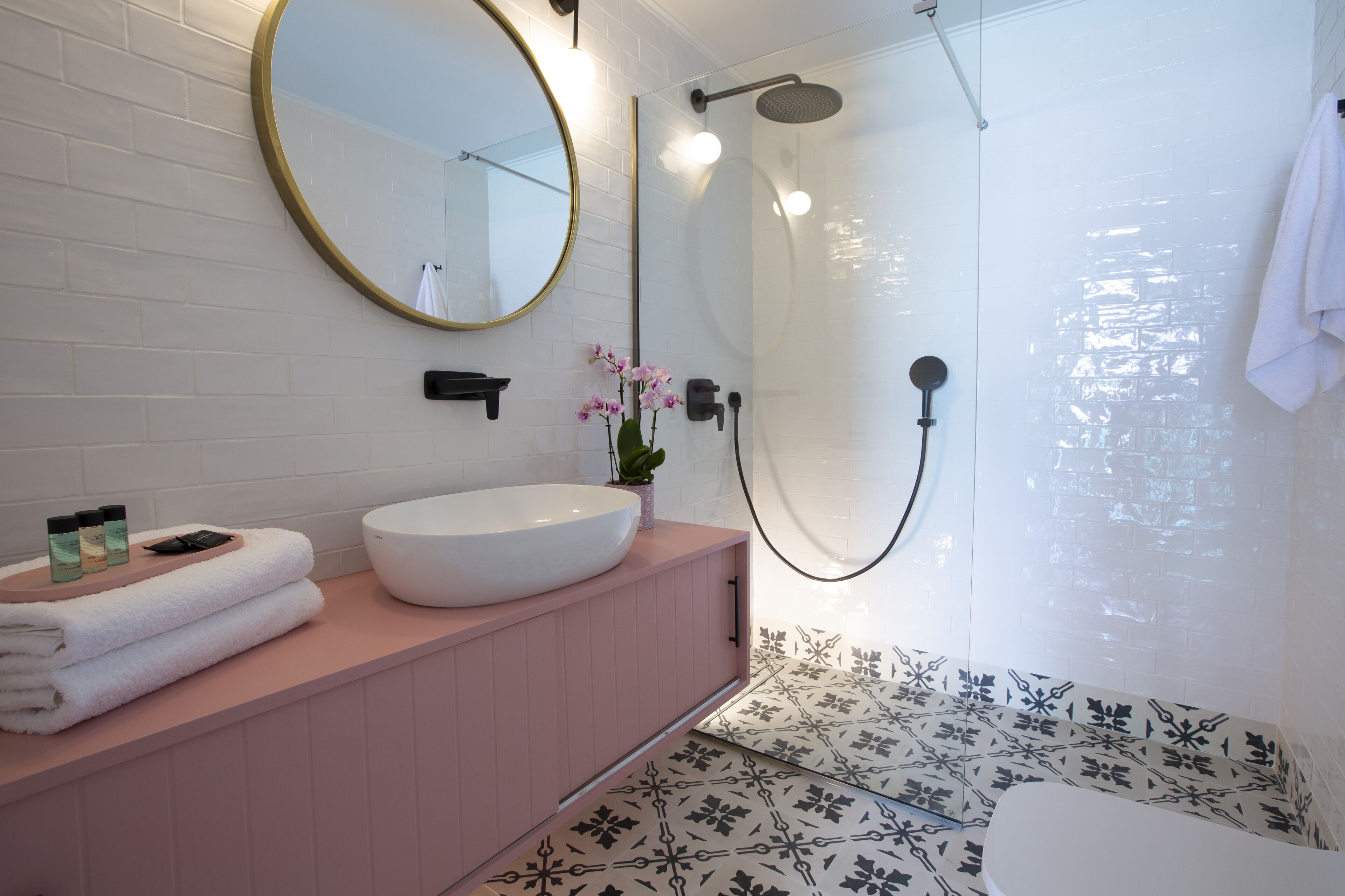 A bathroom with a round mirror, white sink, pink vanity, and a shower with a glass door.