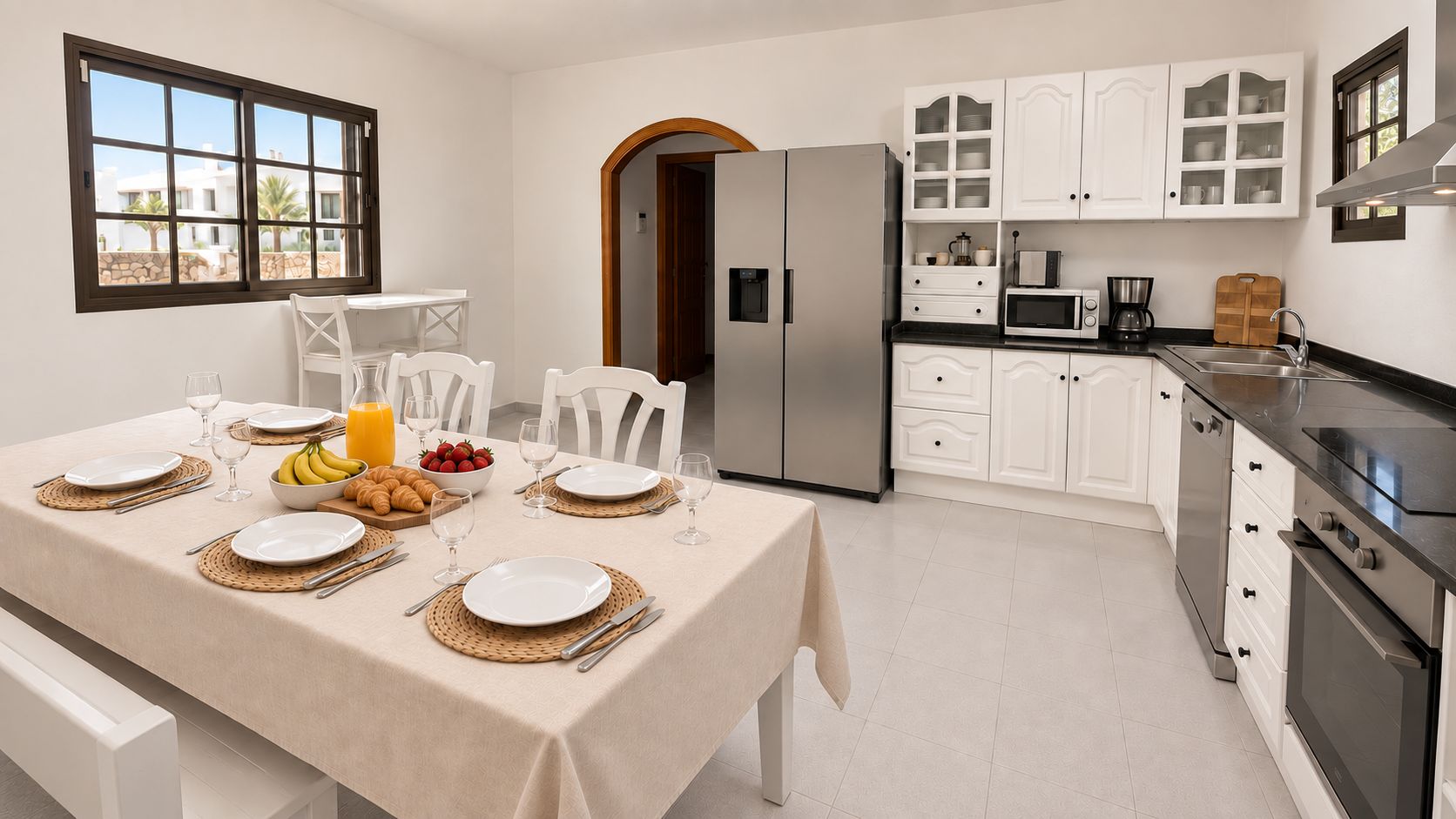 A modern kitchen with white cabinetry, a stainless steel refrigerator, and a dining area set for breakfast with plates, glasses, and croissants.