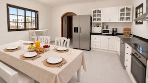 A modern kitchen with white cabinetry, a stainless steel refrigerator, and a dining area set for breakfast with plates, glasses, and croissants.