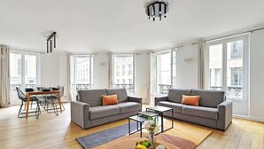 A spacious living room with two grey sofas, a dining table with chairs, and large windows offering a view of buildings. - Renovated and modern 4BD for 12 guests! (Paris)
