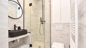 A modern bathroom with a glass-enclosed shower, a round mirror, and a black sink. - Modern 2BD apartment in Parc Belleville (Paris)
